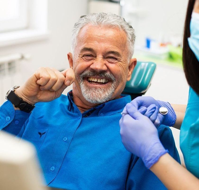 Smiling older man in a blue shirt at a dental appointment. A dental assistant wearing blue gloves holds his hand.