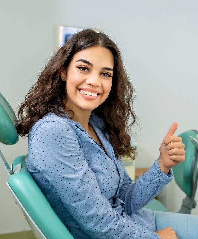 Woman in a blue shirt gives a thumbs up, smiling in a dental chair.