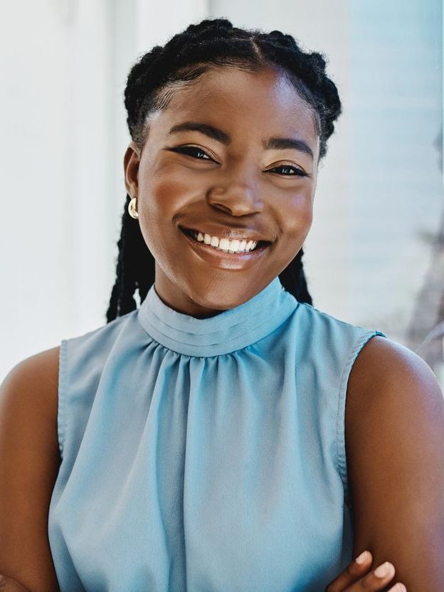 Smiling Black woman in a blue top, braided hair, and gold earrings. She is indoors with a bright background.