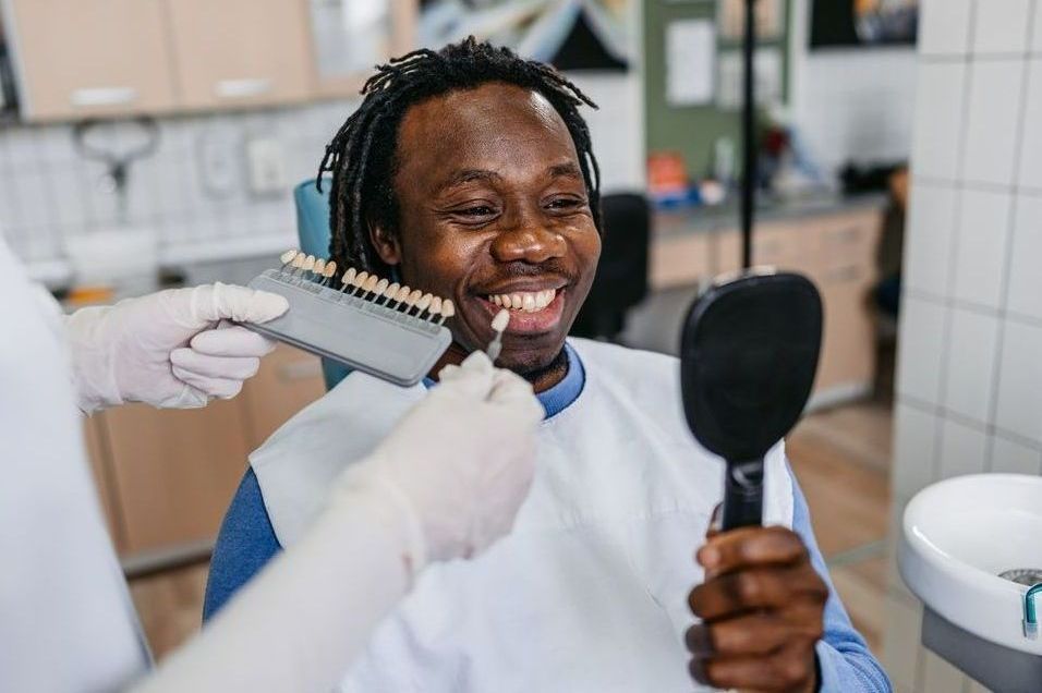 A dentist matching a man's teeth shade with a color chart in a dental office; man smiles while holding a mirror.