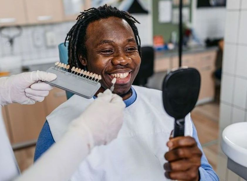 A Black man smiles at the dentist while a shade guide is held to his teeth, selecting a color for dental work.