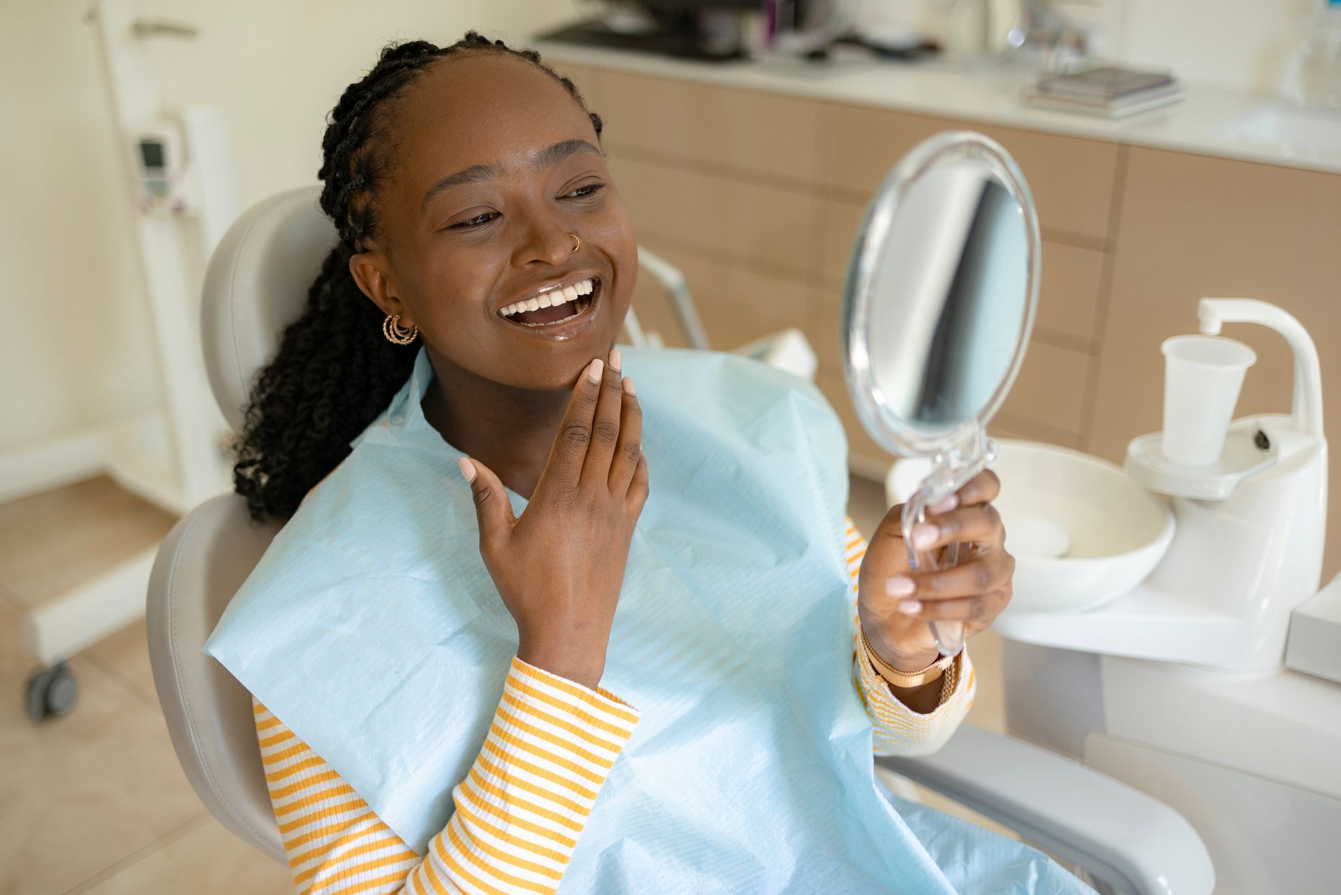 A smiling Black woman in a dentist chair looking at her teeth in a hand mirror, after a dental procedure.