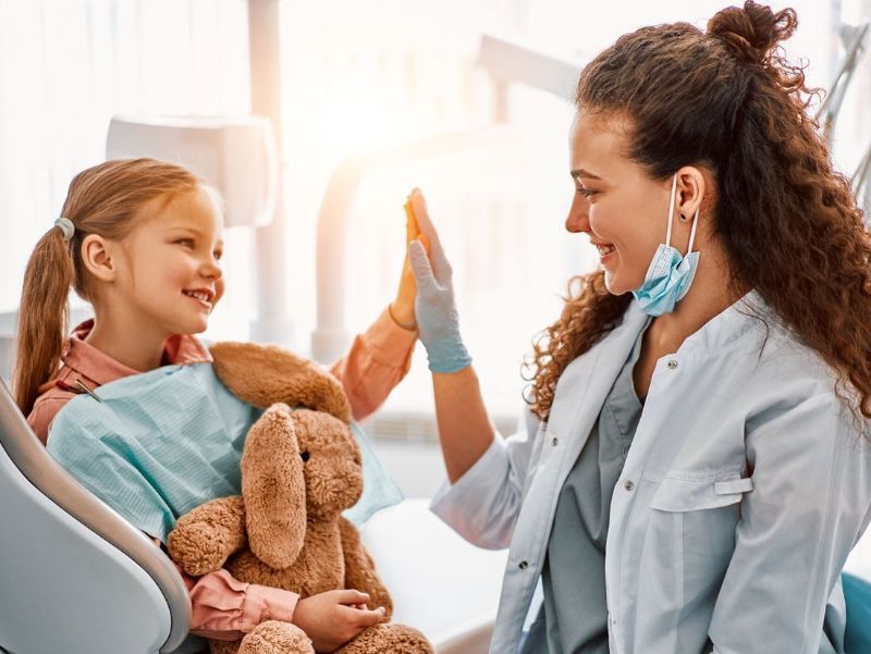 A dentist in a mask and gloves gives a high-five to a smiling young girl in a dental chair, who is holding a stuffed bunny.