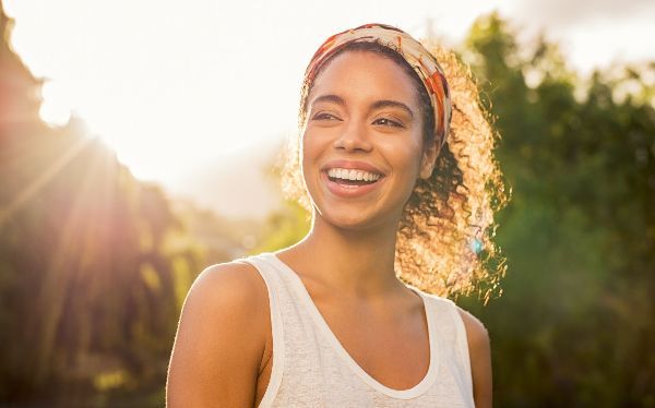 Smiling woman with curly hair and a patterned headband, bathed in sunlight outdoors.