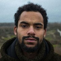 A Black man with a beard and curly hair wearing a dark coat looks directly at the camera in an outdoor setting.