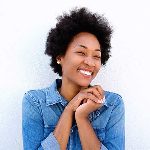 Smiling Black woman with afro, wearing a denim shirt, hands clasped, leaning against a white wall.