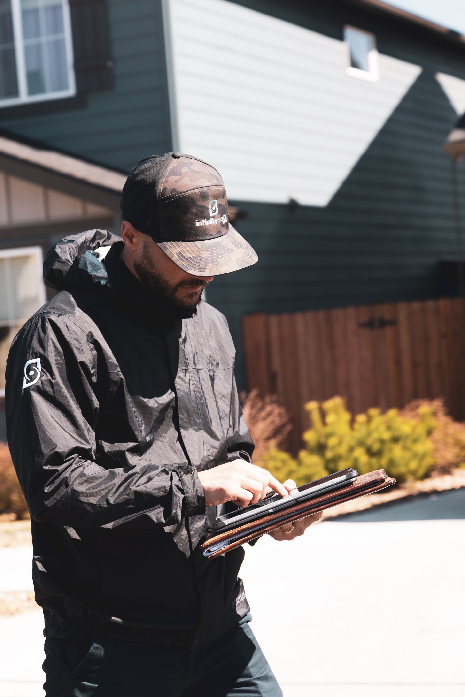 Man in a dark jacket and cap inspecting a tablet outdoors in front of a house and fence.