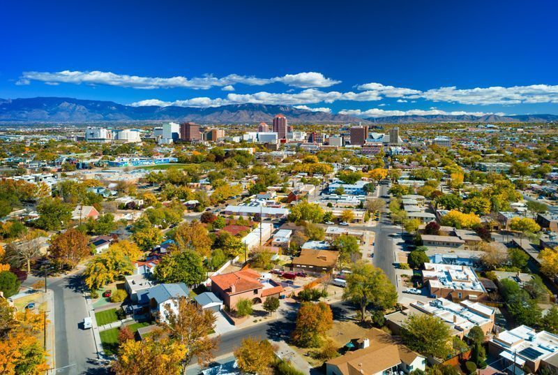 An aerial view of a city with mountains in the background. Aerial view of a leafy suburban neighborhood with city skyline and mountains under a bright blue sky.