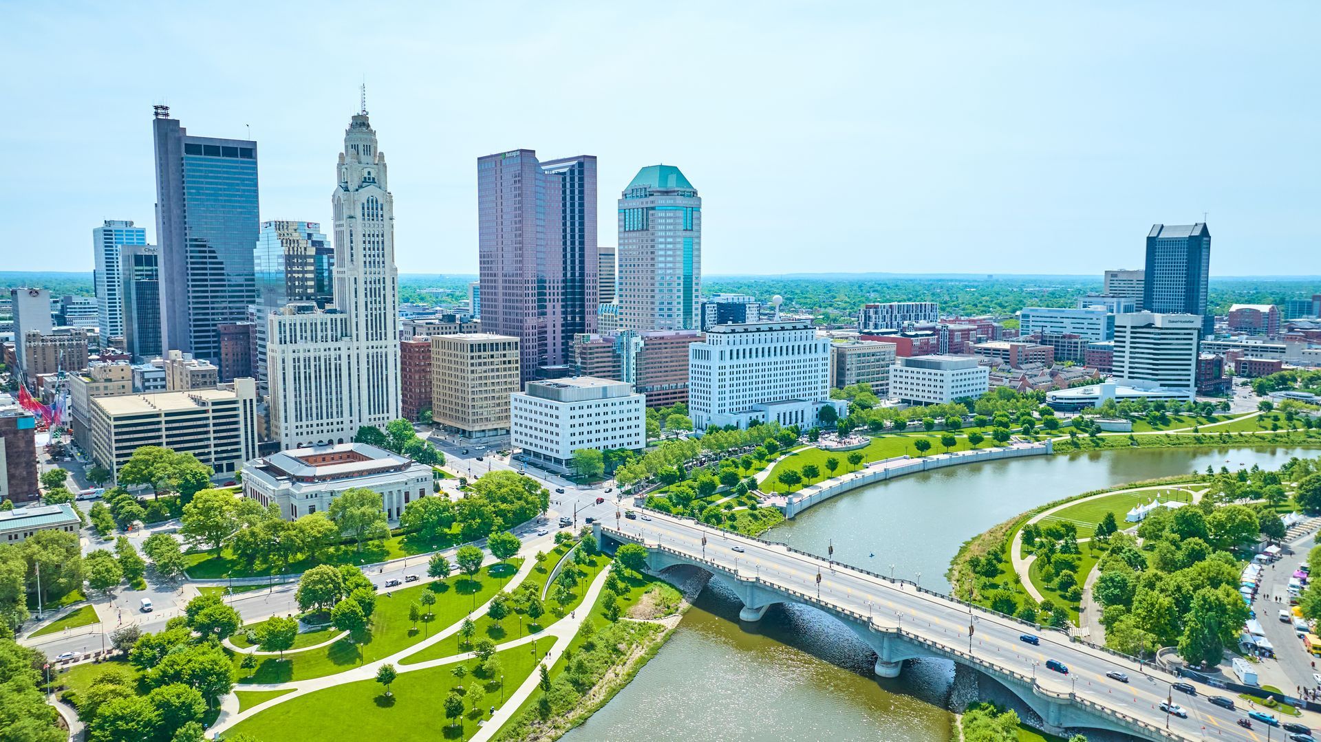 Aerial cityscape with modern high-rises, a winding river, bridge, and green parks under a clear sky.