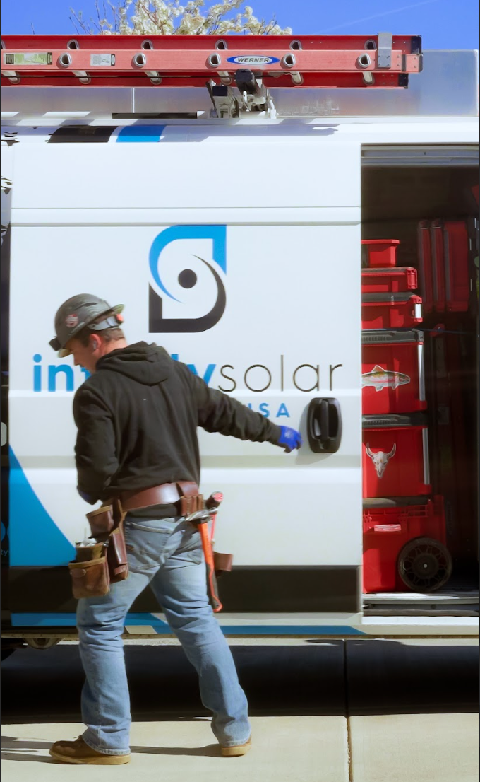 Worker in hard hat walking beside a white utility van with open side compartment and red ladder on top