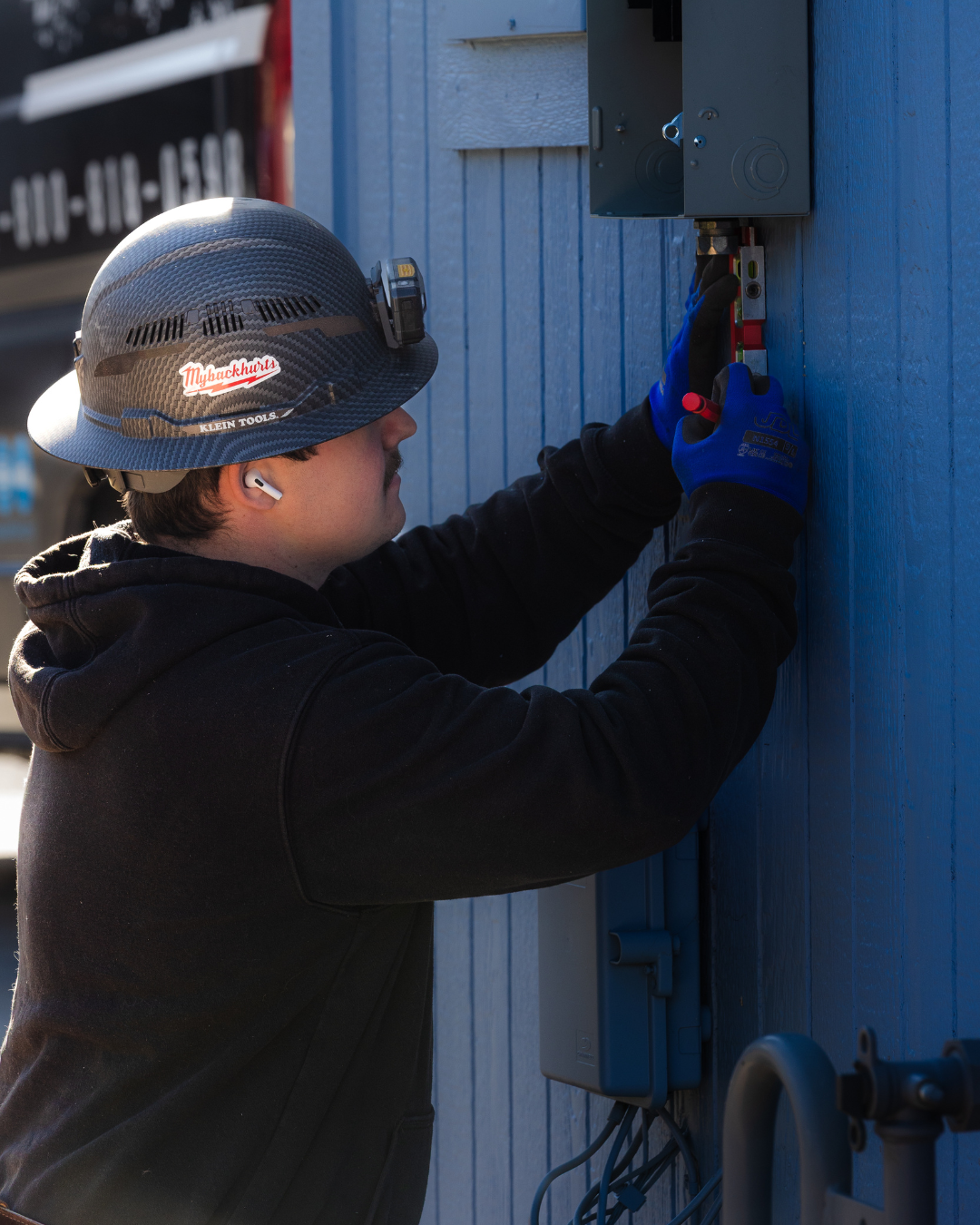 Worker in hard hat and gloves adjusting an electrical panel on a blue building exterior.