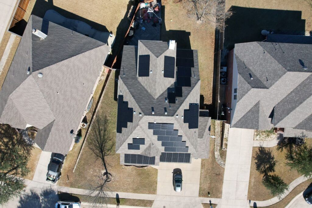 An aerial view of a house with solar panels on the roof Aerial view of three suburban houses with dark gray roofs and parked cars on driveways.