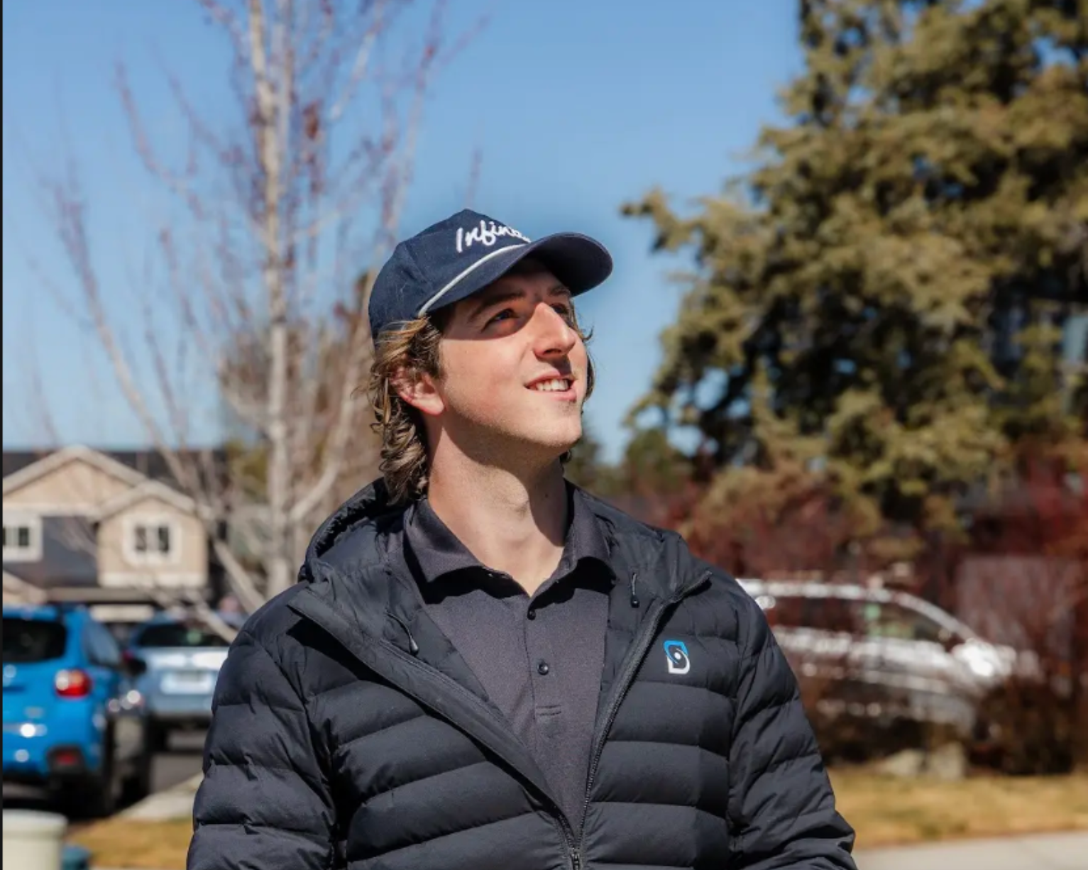 Person in a dark jacket and cap smiling outdoors on a sunny residential street.