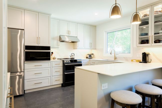 Modern white kitchen with stainless steel appliances, breakfast bar with stools, and brass pendant lights.