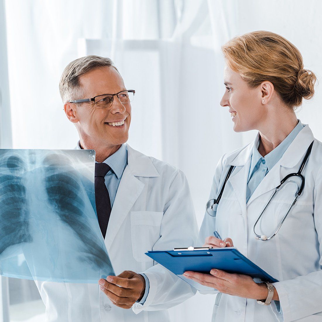 Doctors examine chest X-ray in a bright room. One holds the X-ray, the other holds a clipboard, both smiling.