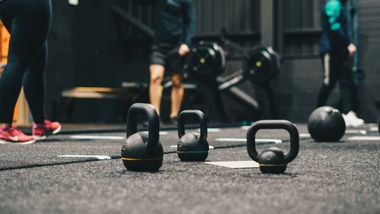 Kettlebells on gym floor; people in workout attire. Dark interior, training area.