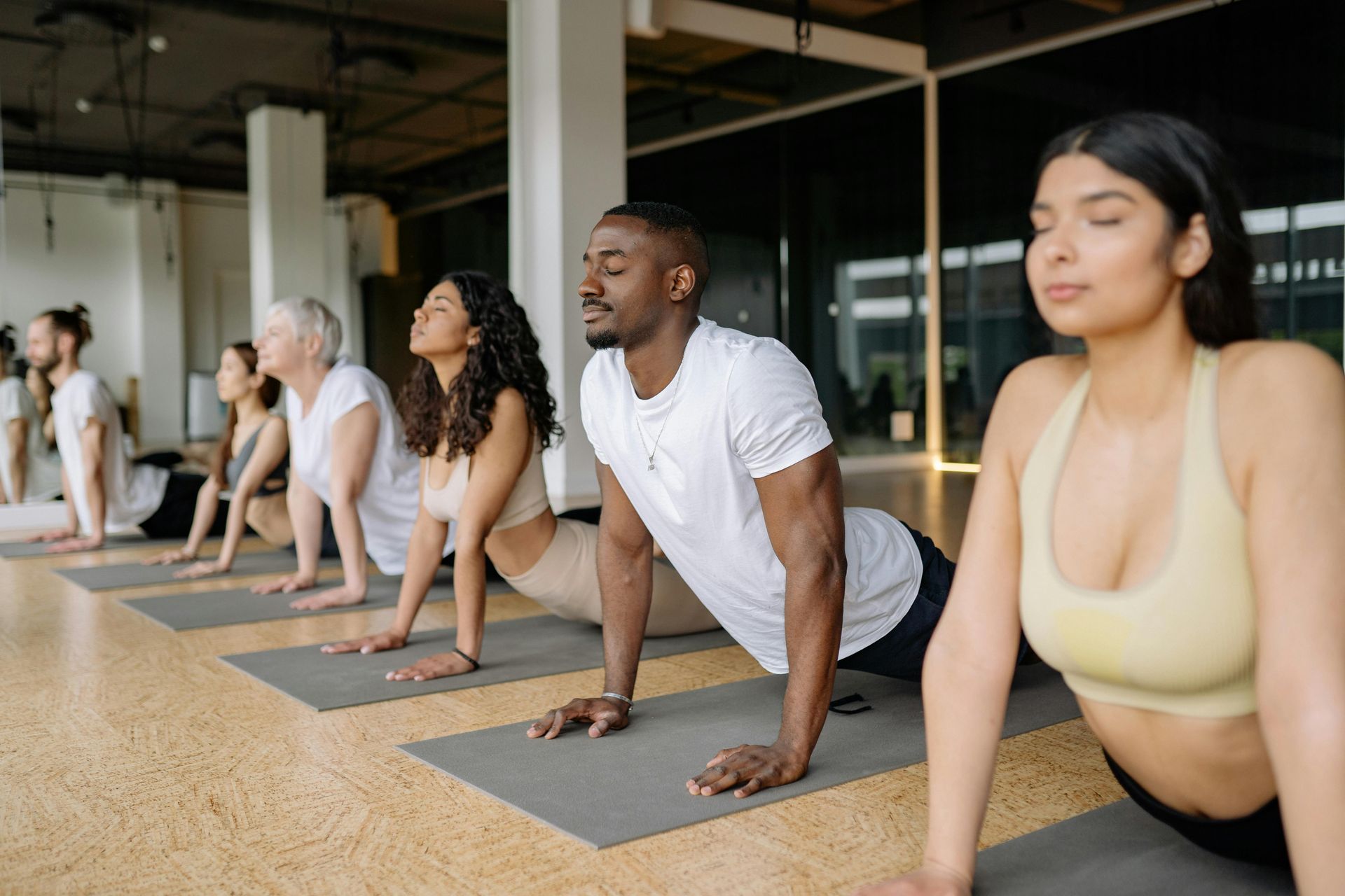 People in yoga class doing upward-facing dog pose. Diverse group with eyes closed, on mats. Bright indoor setting.