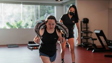Woman doing a weighted carry exercise with trainer in a gym.