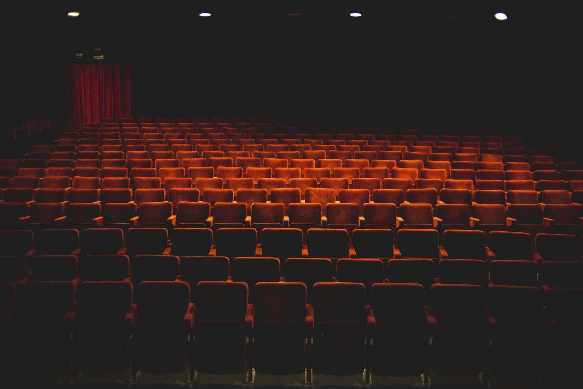 Empty theater auditorium with rows of red seats, stage, and red curtain. Dark lighting.