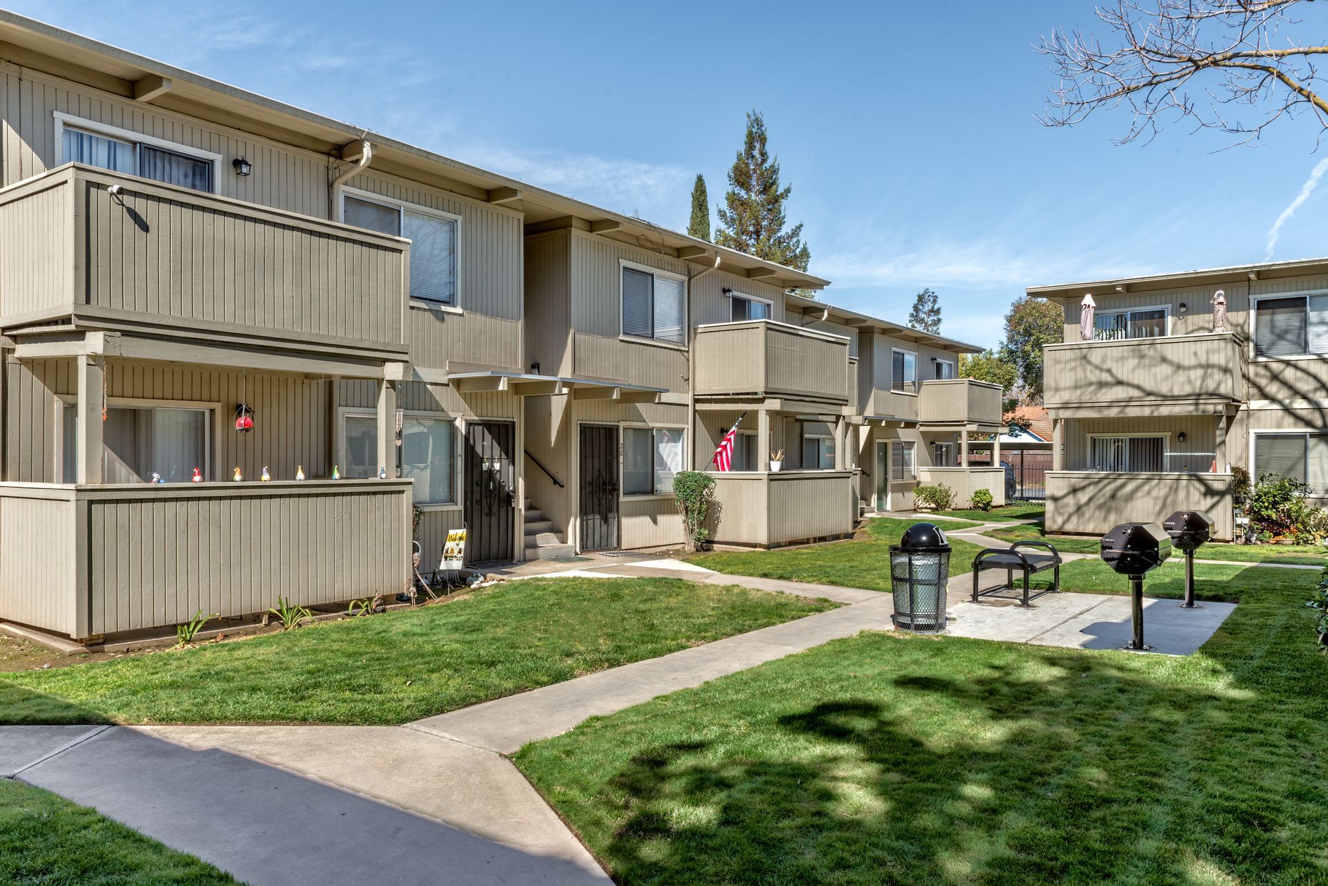 Apartment buildings with balconies, green grass, and a barbecue area on a sunny day.