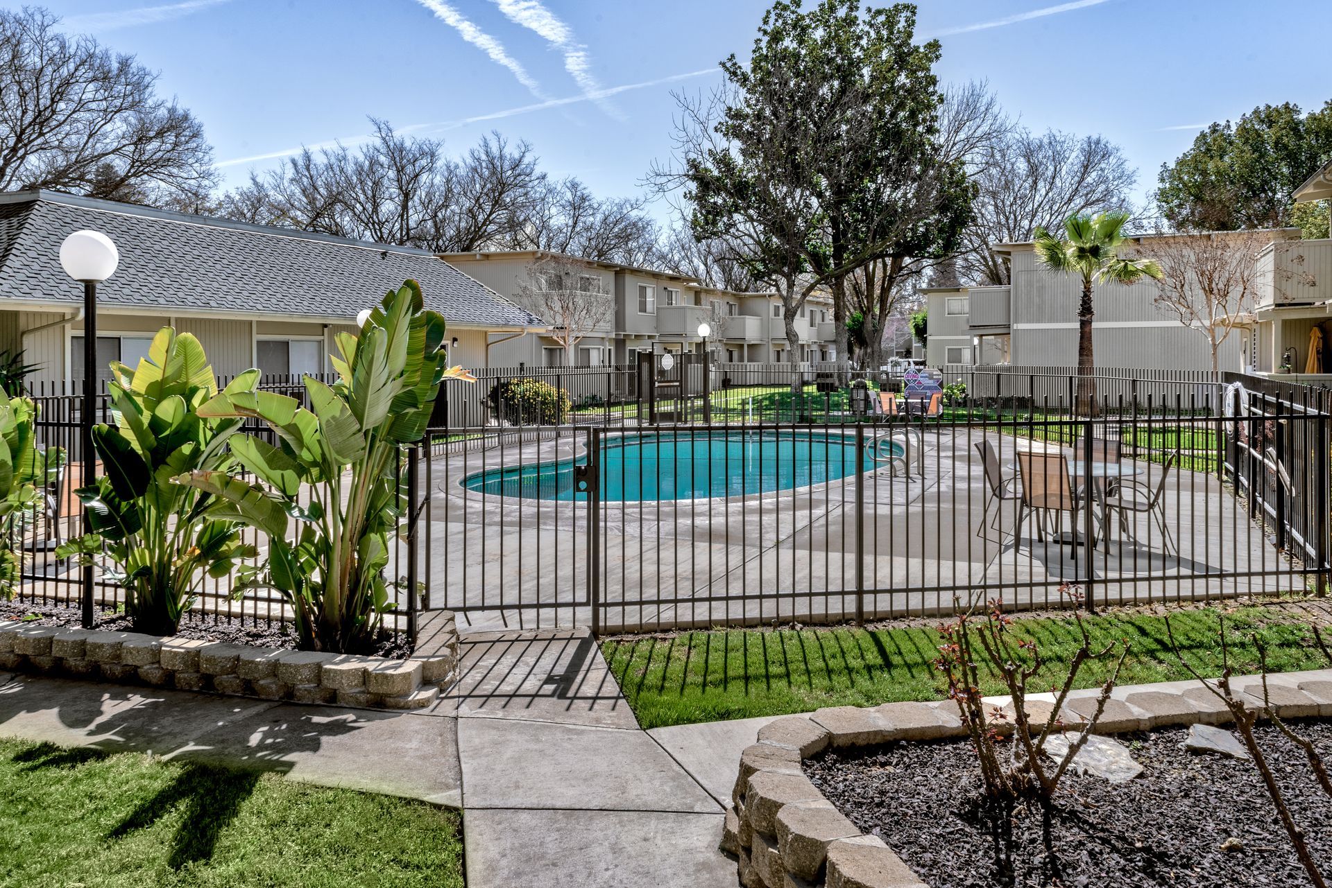 Swimming pool surrounded by a black fence in a residential area. Green grass, trees, and buildings in the background.