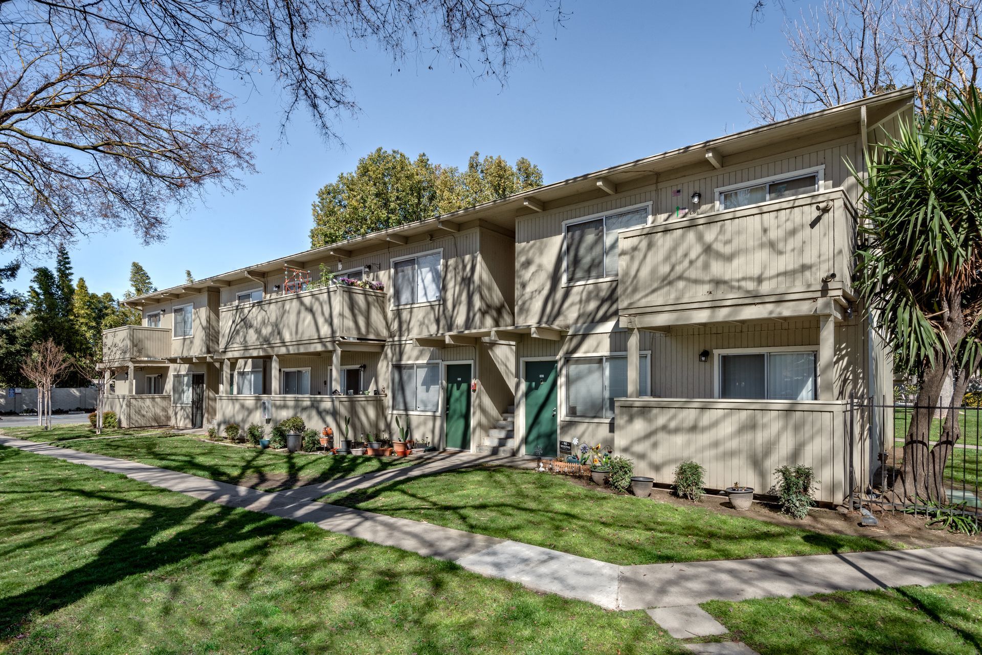 Two-story beige apartment complex with balconies, green doors, and a grassy yard under a blue sky.