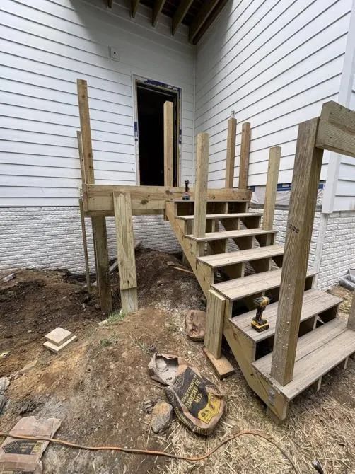 A partially constructed wooden staircase leading to a doorway on the side of a white house, surrounded by dirt ground.