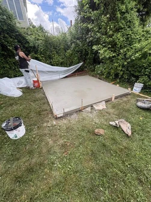 A person pulls a large plastic sheet over a freshly poured concrete slab in a grassy backyard.