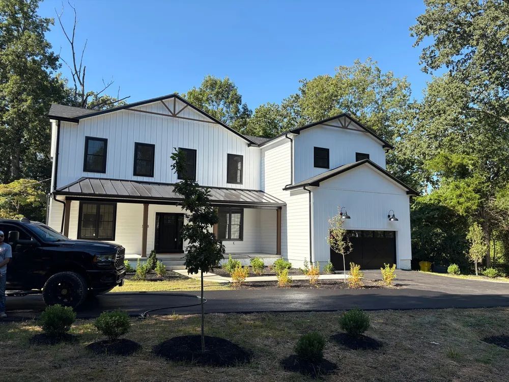 A white two-story modern farmhouse with black window frames and a dark garage, situated in a wooded area with a driveway.