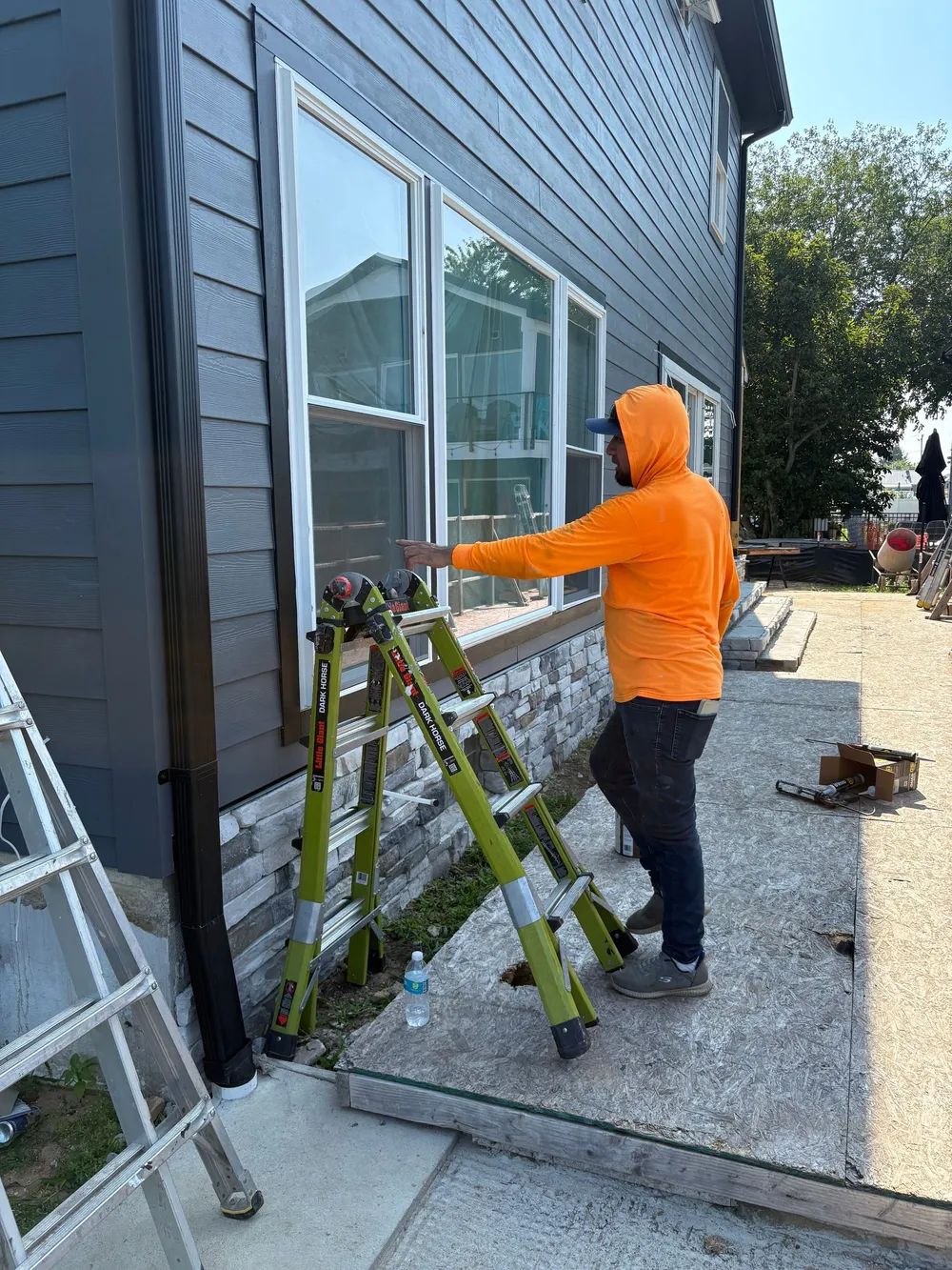 A person in an orange hooded sweatshirt works on a window frame exterior on a house with dark siding and stone accents.