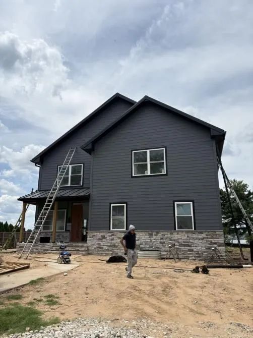 A person stands in front of a partially constructed two-story house with dark gray siding and a stone base foundation.