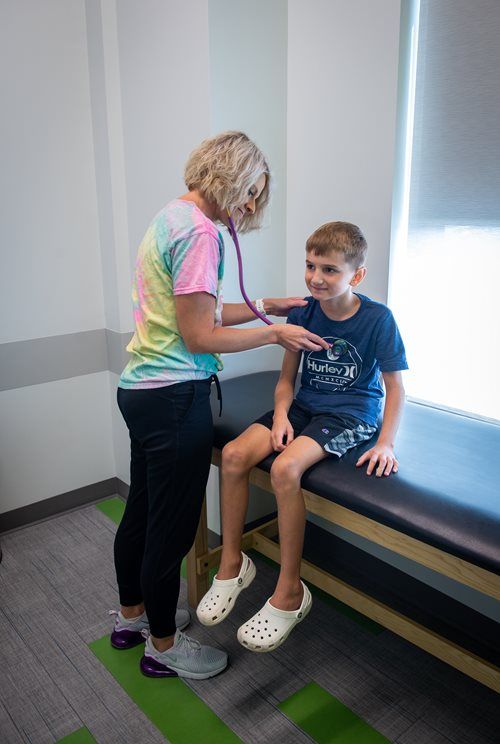 A clinician in a tie-dye shirt uses a stethoscope to examine a patient sitting on an exam table in a clinical room.