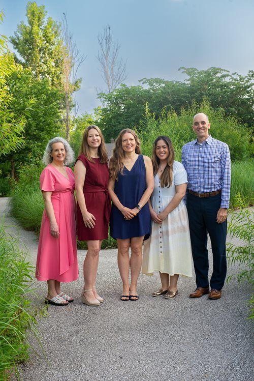 Five people stand side-by-side on a gravel path in a lush, green garden, smiling at the camera.