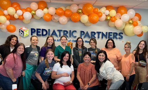 A large group of smiling staff stands in front of a Pediatric Partners sign decorated with orange and white balloons.