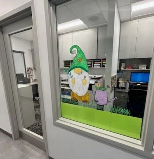 A gnome drawing with a purple watering can displayed on an office glass window with green trim.