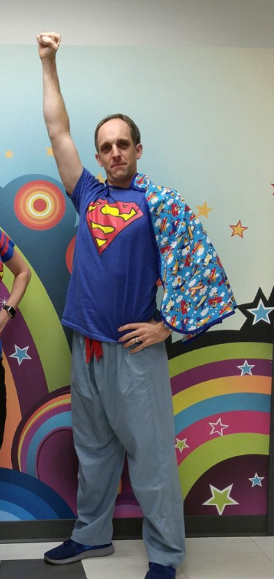 A person in a Superman t-shirt and patterned cape strikes a hero pose in front of a colorful wall with stars and rainbows.