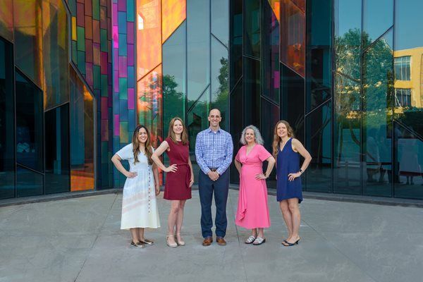 Five people stand in a line posing for a photo in front of a modern, multi-colored glass building facade.