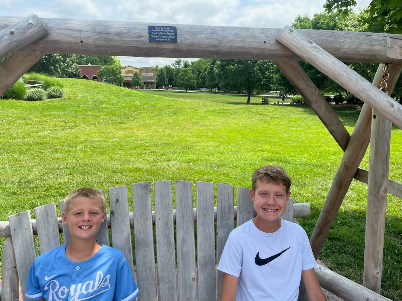 Two smiling children seated on a wooden swing bench in a grassy park under a rustic archway