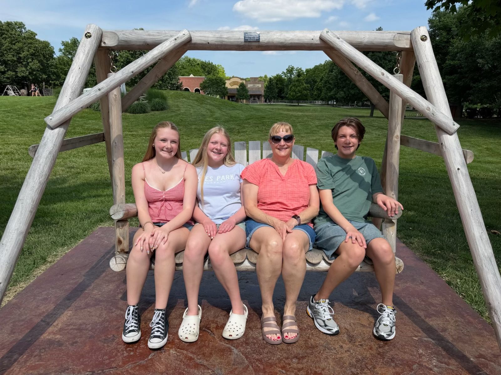 Four people sitting on a wooden swing bench in a grassy park, smiling at the camera.