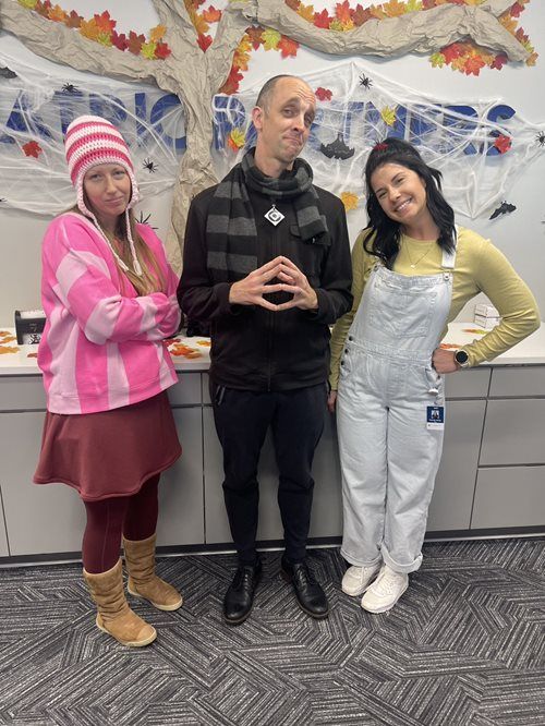 Three people stand side-by-side in costumes in front of a decorative fall-themed wall.