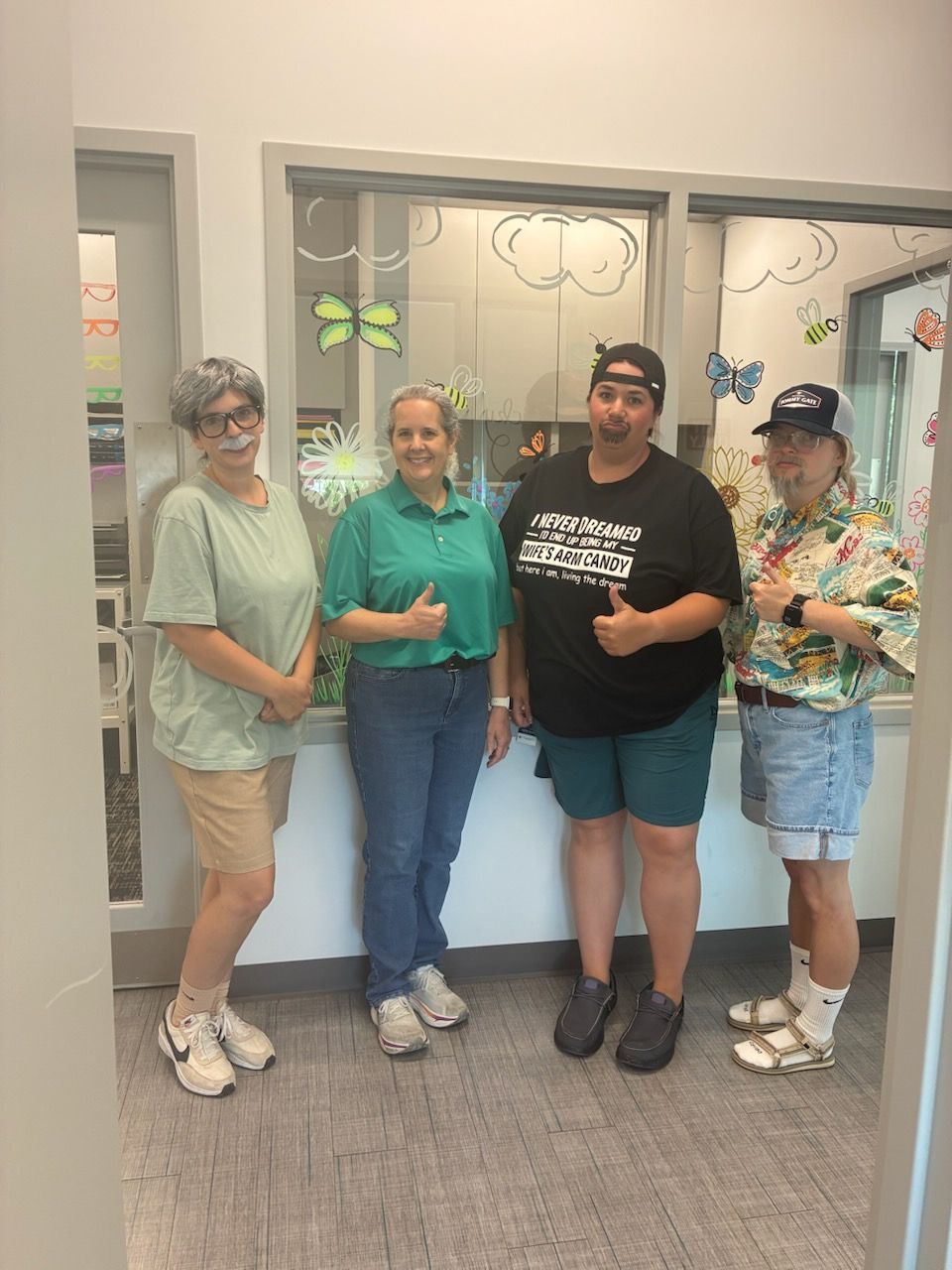 Four female staff dressed as their fathers smiling and posing in front of a windowed wall.
