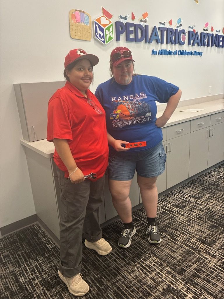 Two people posing at our check in desk, one in Chiefs hat and shirt and one in a KU basketball shirt holding a carpenter's level.
