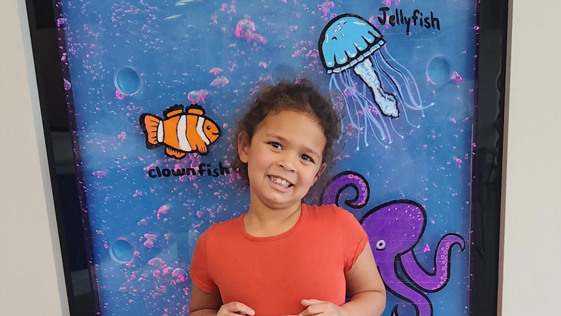Smiling child in orange shirt standing before a colorful underwater-themed display in Olathe with fish and a jellyfish