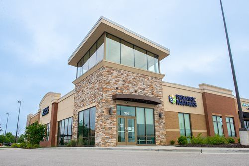 A modern, tan-brick commercial building with a central stone tower and glass-fronted storefronts under a cloudy sky.