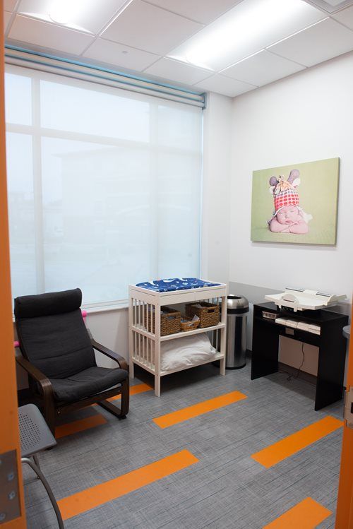 A nursing or changing room with a black armchair, a baby changing table with baskets, a side table, and a wall painting.