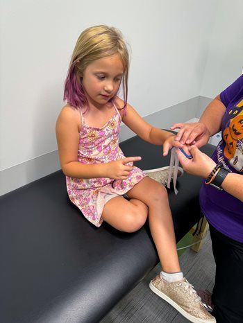 A person in a medical setting bandages the finger of a child wearing a floral dress.
