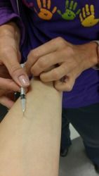 A person wearing a purple shirt with colorful hand prints holds a small syringe while injecting it into an arm.