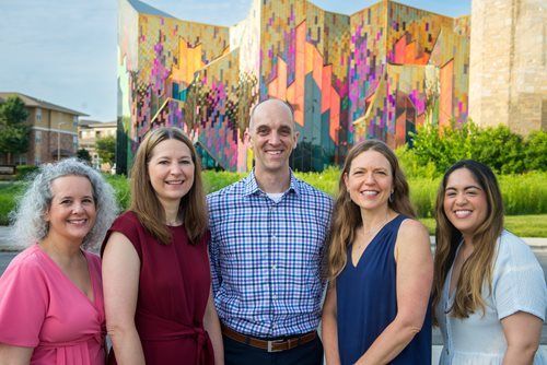 Five people pose side-by-side in front of a colorful, geometric outdoor mural on a sunny day.