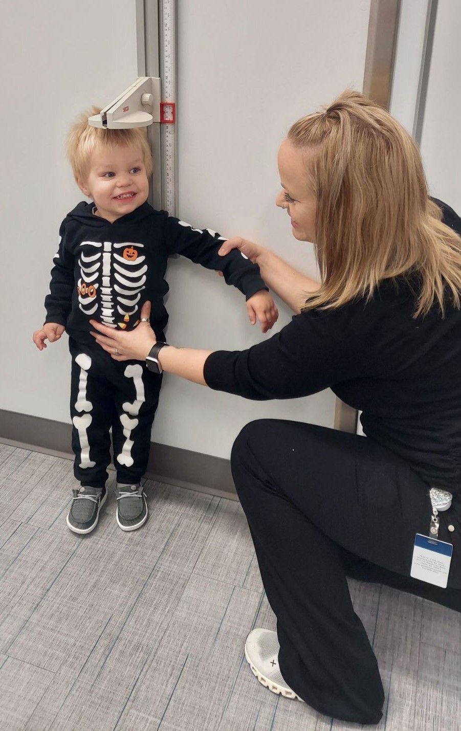 A healthcare worker measures the height of a child wearing a skeleton costume standing against a wall-mounted stadiometer.