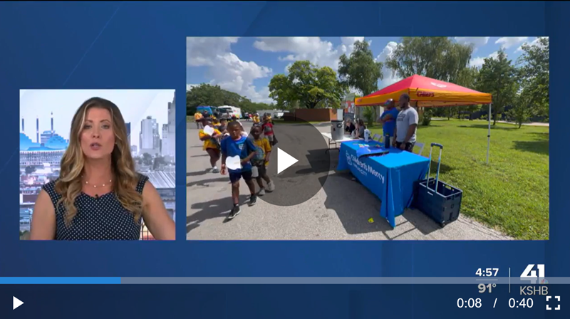 News anchor beside outdoor event setup with blue table, orange canopy, and people walking on a sunny path
